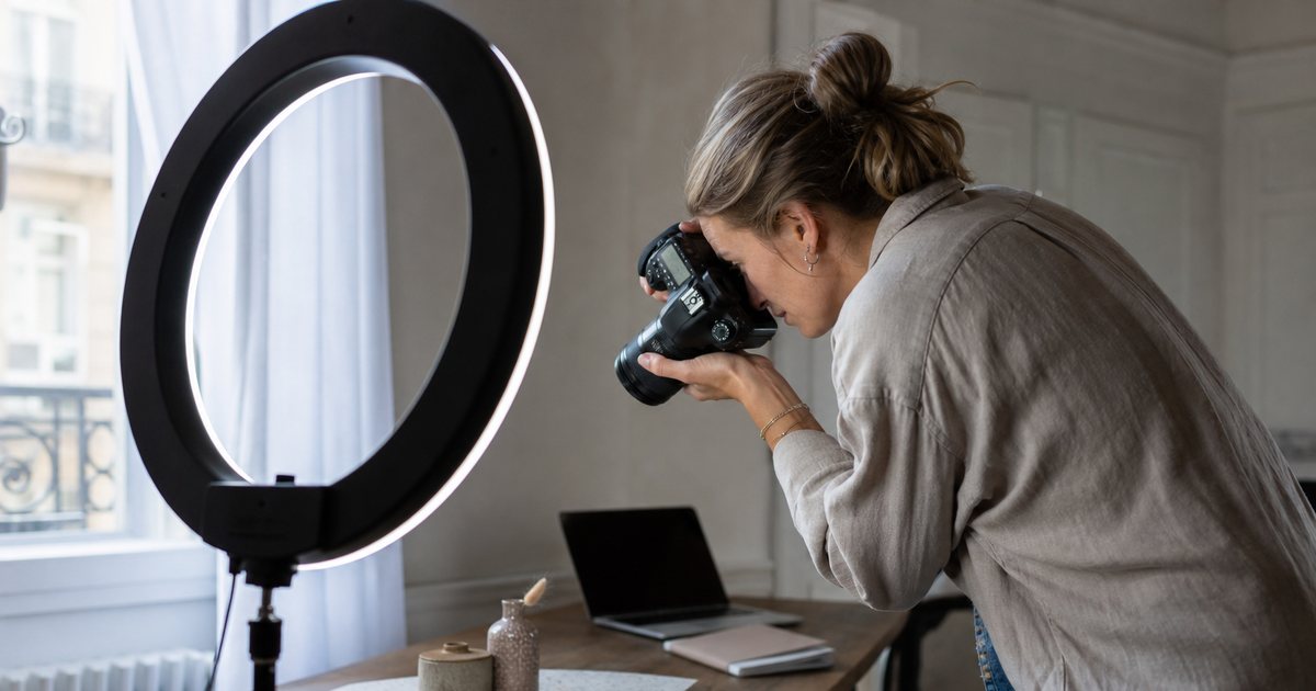 Content creator photographing a product on a tabletop with a ring light in a home studio, laptop and notebook to the side, natural light from a window