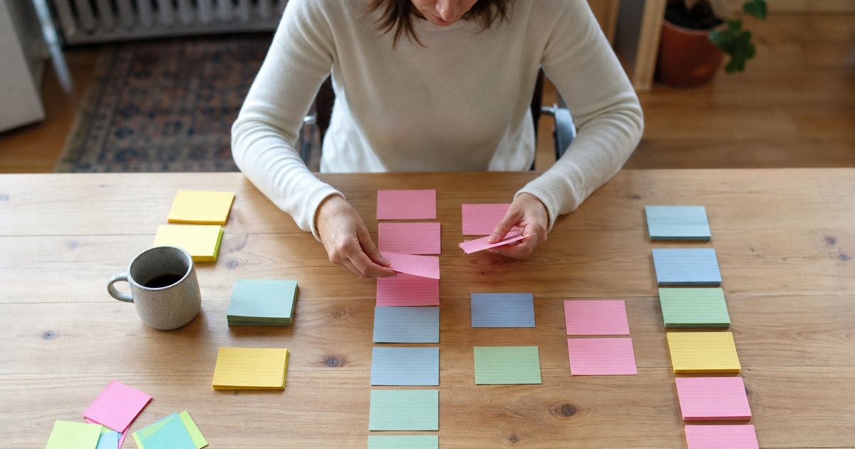 Person sorting colored index cards into neat stacks on a large wooden table, sticky notes and a mug nearby, overhead view in soft daylight