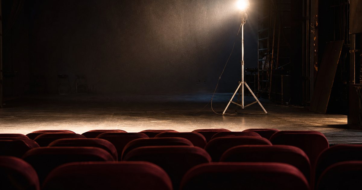 An empty theater stage lit by a single bare work light on a metal stand, rows of empty red seats in the foreground, dust visible in the beam